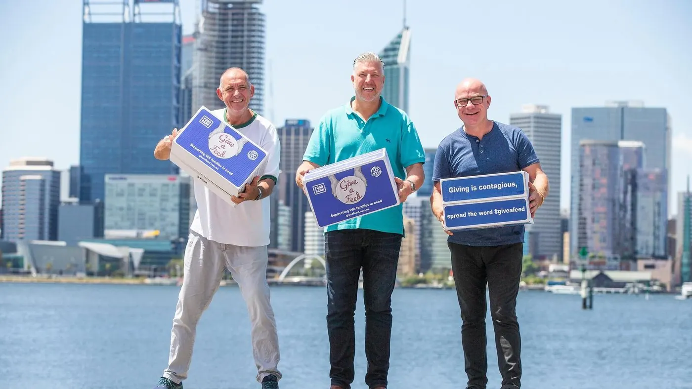 Three Give A Feed supporters holding hamper boxes on the Swan River foreshore with the Perth city skyline behind, one box reading Giving is contagious, spread the word #giveafeed
